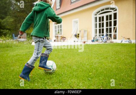 Rear view of boy wearing wellingtons playing football in the garden Stock Photo