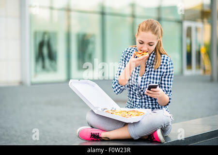 Teenager eating pizza in street and browsing internet on phone Stock Photo