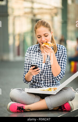 Teenager eating pizza in street and browsing internet on phone Stock Photo