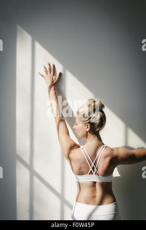 A blonde woman standing in front of a white wall, doing yoga, her arm raised, touching the wall. Stock Photo