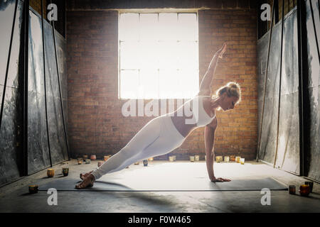 A blonde woman, in a white crop top and leggings, standing on a yoga mat surrounded by candles, doing yoga, her arm raised. Stock Photo