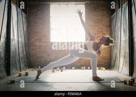 A blonde woman, in a white crop top and leggings, standing on a yoga mat surrounded by candles, doing yoga, her arm raised. Stock Photo