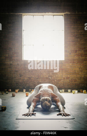 A blonde woman in a white crop top and leggings, bending down on a yoga mat surrounded by candles, doing yoga. Stock Photo