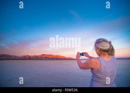 Woman taking smartphone photograph of salt flats and distant mountains at sunset, Calico, California, USA Stock Photo