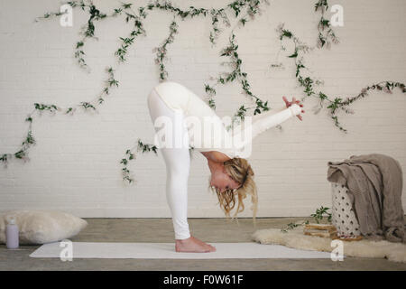 A blonde woman in a white leotard and leggings bending down, stretching. A creeper plant on the wall behind her. Stock Photo