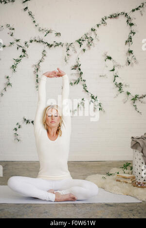 A blonde woman sitting on a white mat in a room stretching her arms. A creeper plant on the wall behind her. Stock Photo
