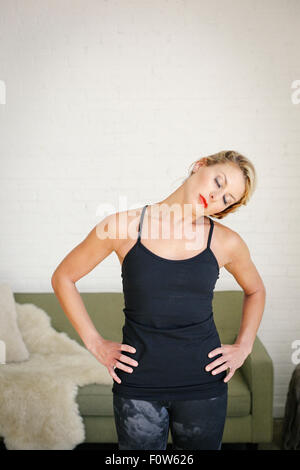 A blonde woman in a black leotard standing in a room, doing yoga, her hands on her hips, stretching her neck.