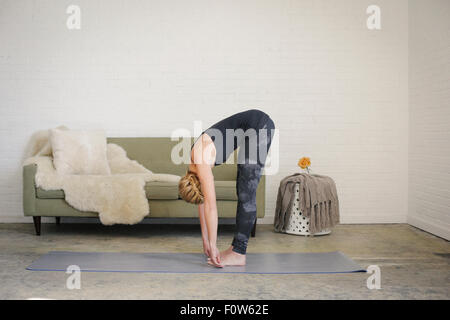 A blonde woman standing on a yoga mat in a room, doing yoga, bending down, touching her toes with her hands. Stock Photo