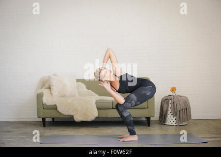 A blonde woman in a black leotard and leggings, standing on a yoga mat in a room, doing yoga, squatting down with her arms behind her head. Stock Photo