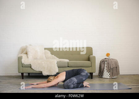 A blonde woman kneeling on a yoga mat in a room, doing yoga, her arm stretched out in front of her. Stock Photo