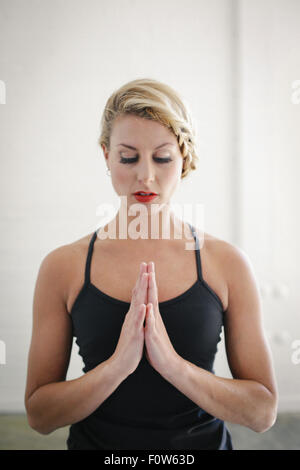 A blonde woman in a black leotard doing yoga, standing with her eyes closed and her hands together.