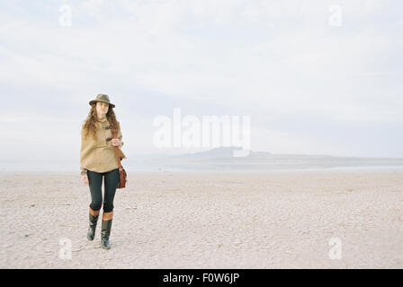 A woman with brown, long and curly hair walking through a desert plain, wearing a hat and carrying a leather bag. Stock Photo