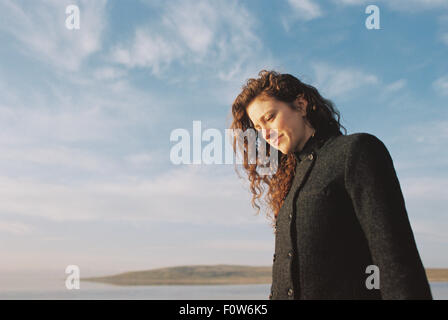 Woman with brown, long and curly hair standing on the shore of a lake. Stock Photo