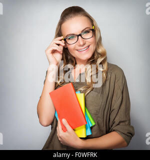Portrait of cute blond student girl over gray background, wearing glasses and holding in hands colorful books, back to school Stock Photo