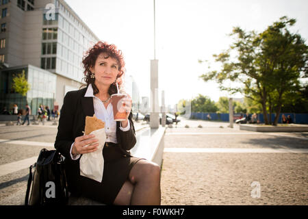 Senior business woman on fast food lunch - drinking take-away coffee and eating sandwich in street Stock Photo