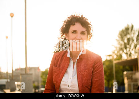 Portrait of smiling senior woman wearing red jacket in street with sun in background Stock Photo
