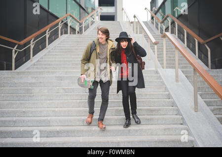 Young couple of caucasian woman and man, with skate and moustache, walking down the stairs hugging and smiling - youth, carefree, freshness concept Stock Photo