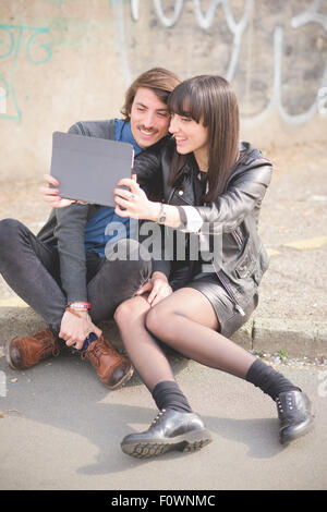 Couple of young beautiful caucasian man and woman seated on a sidewalk in the city using a tablet taking a selfie - social network, technology, communication concept Stock Photo