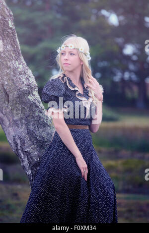 a blonde girl in a floral dressl is leaning against a tree Stock Photo
