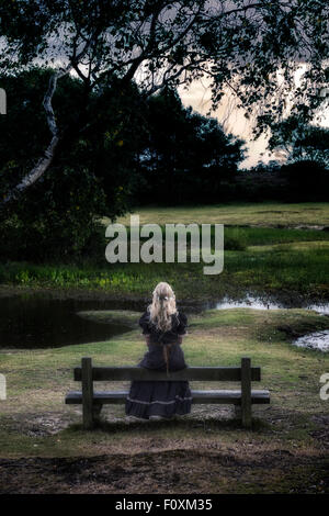 a blonde girl in a floral dressl is sitting on a bench at a pond Stock Photo