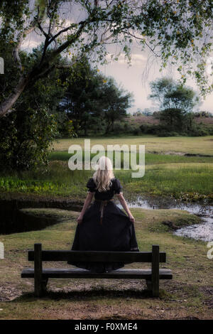 a blonde girl in a floral dressl is standing on a bench at a pond Stock Photo