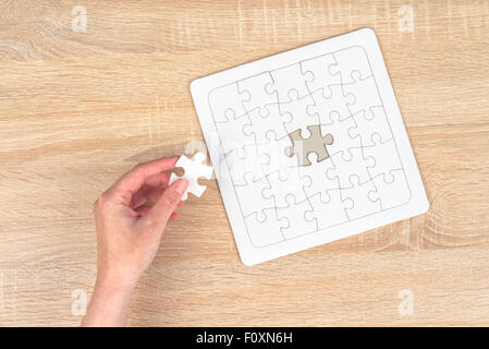 Female hand putting a missing piece and solving blank white jigsaw puzzle placed on top of old wooden oak table, top view Stock Photo