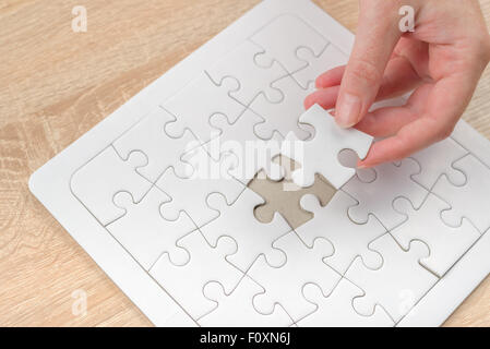 Female hand putting a missing piece and solving blank white jigsaw puzzle placed on top of old wooden oak table, close up Stock Photo