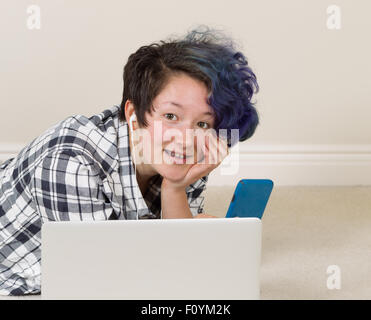 Smiling teen girl, looking forward, with cell phone in hand with computer in forefront while lying down listening to music at ho Stock Photo