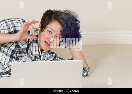 Smiling teen girl, looking forward, giving okay sign while using computer and listening to music at home. Stock Photo