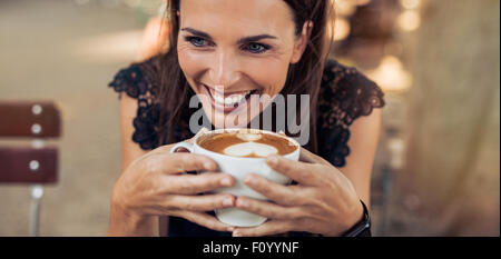 Close up shot of cheerful young woman drinking coffee at a cafe and looking away. Caucasian female enjoying a cup of coffee. Stock Photo