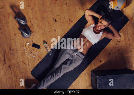 Overhead view of smiling female working out at the gym. Muscular young woman lying on exercise mat with her hands behind head do Stock Photo
