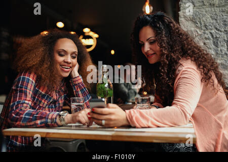 Portrait of  young women sitting at a cafe using mobile phone. Two young girls reading a text message on mobile phone. Stock Photo