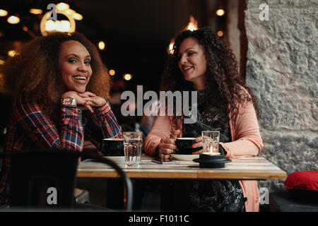 Two women sitting at a restaurant smiling. Young friends in a cafe, with one looking at camera smiling. Stock Photo