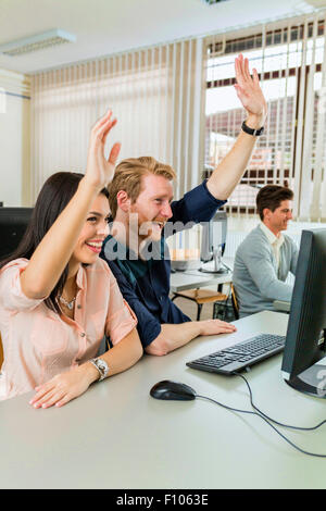 Beautiful young woman and a man raising hands in classroom in  while sitting in fron of a desktop Stock Photo