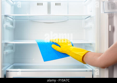 Woman's hand in yellow rubber protective glove cleaning white open empty refrigerator with blue rag Stock Photo