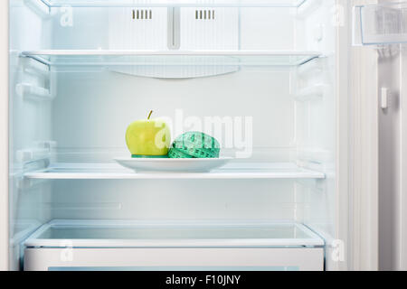 Green apple with measuring tape on white plate in open empty refrigerator. Weight loss diet concept. Stock Photo