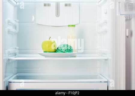Green apple with measuring tape on white plate and a glass bottle of yogurt in open empty refrigerator. Weight loss diet concept Stock Photo