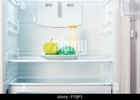 Green apple with measuring tape on white plate and a glass bottle of yogurt in open empty refrigerator. Weight loss diet concept Stock Photo