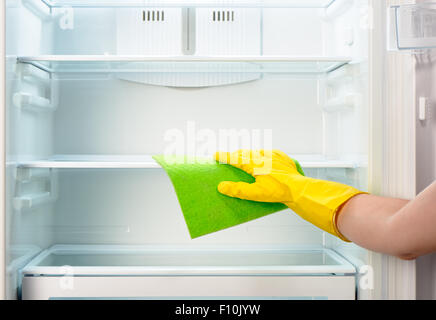 Woman's hand in yellow rubber protective glove cleaning white open empty refrigerator with green rag Stock Photo