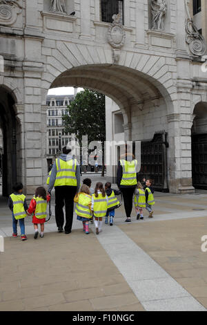 Nursery school children & child minders wearing high visibility fluorescent safety jackets hold hands on walk in a street in London UK   KATHY DEWITT Stock Photo