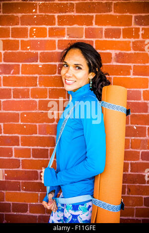Hispanic woman carrying yoga mat at brick wall Stock Photo