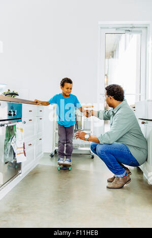 Father helping son balance on skateboard in kitchen Stock Photo