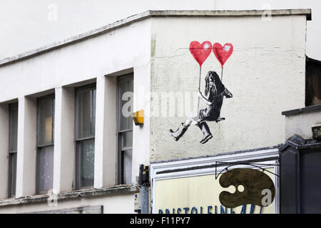 Street art, Banksy style, in Park Row Bristol. The image shows a girl on a asking that is suspended by two heart shape balloons. The artist is jps Stock Photo
