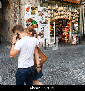 Young couple hugs while man talks on mobile phone and both walk in front of grocery store in the old district of Naples Stock Photo