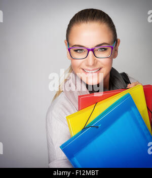 Portrait of beautiful smart student girl wearing glasses, standing with colorful folders over gray background Stock Photo