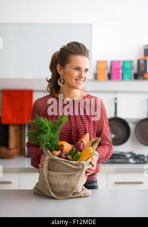 An elegant woman is standing, smiling and looking into the distance. She is holding a burlap sac filled with a variety of fall fruits and vegetables. The colours and flavours of the autumn abound. Stock Photo