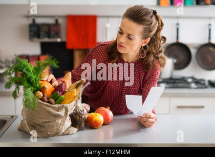 An elegant woman is leaning on the kitchen counter, checking the contents of her burlap bag which is filled with fresh autumn fruits and vegetables. She is double-checking that she has everything. Stock Photo