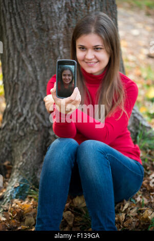 Pretty young woman sitting near tree and taking selfie with mobile phone in the autumn park Stock Photo