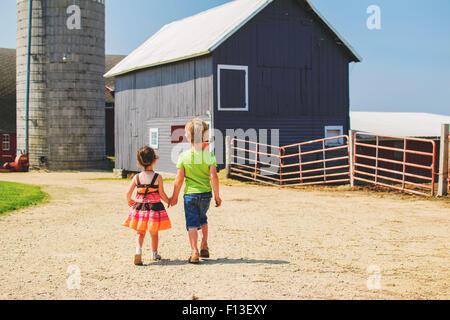 Rear view of a girl and boy holding hands, walking towards farm buildings, USA Stock Photo