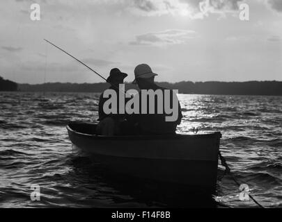 1930s TWO ANONYMOUS MEN WEARING HATS IN ROWBOAT FISHING SILHOUETTED AT SUNRISE Stock Photo
